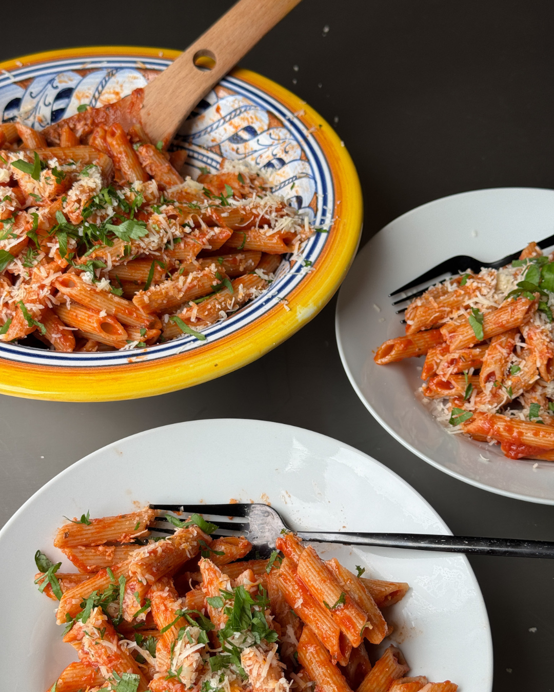 Two plates of penne pasta with a red sauce and herbs, one on a yellow plate with a wooden spoon, on a dark surface.