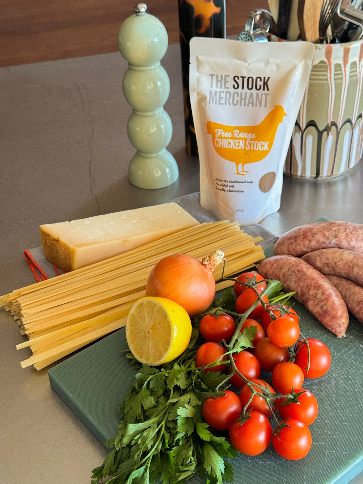 Ingredients for a pasta dish including spaghetti, chicken stock, tomatoes, lemon, and herbs on a kitchen counter.