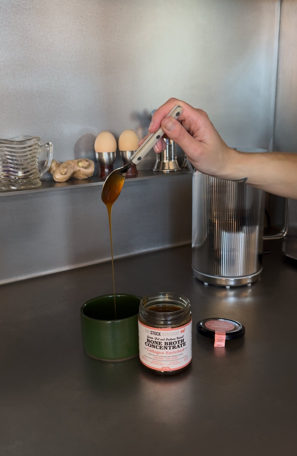 Hand pouring a bone broth concentrate from a spoon into a mug on a kitchen counter.